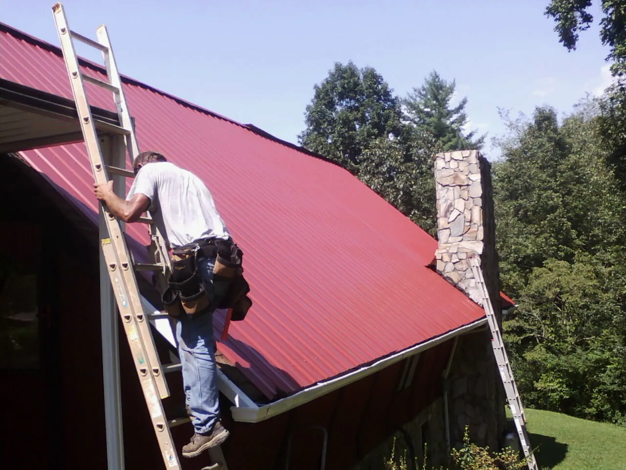 Roofer on a ladder beside a red metal roof and stone chimney — Soffit & Fascia Repair by Roofing Brothers in Gonzales